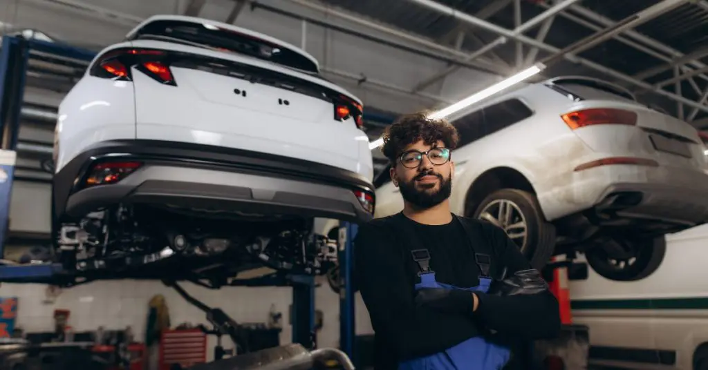 Payment assist car repair payment plan: A young male mechanic with curly hair, glasses and a beard stands with arms folded in a garage workshop, with two white cars raised on hydraulic lifts behind him.
