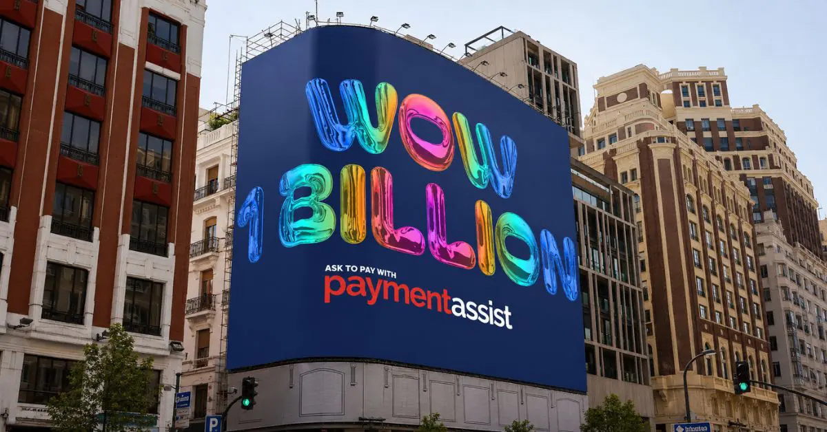 Payment assist passes 1bn customer advances: A large digital billboard on the side of a city centre building displaying the text "WOW 1 BILLION" in colourful metallic balloon-style lettering, with the Payment Assist logo and "Ask to pay with" tagline below.