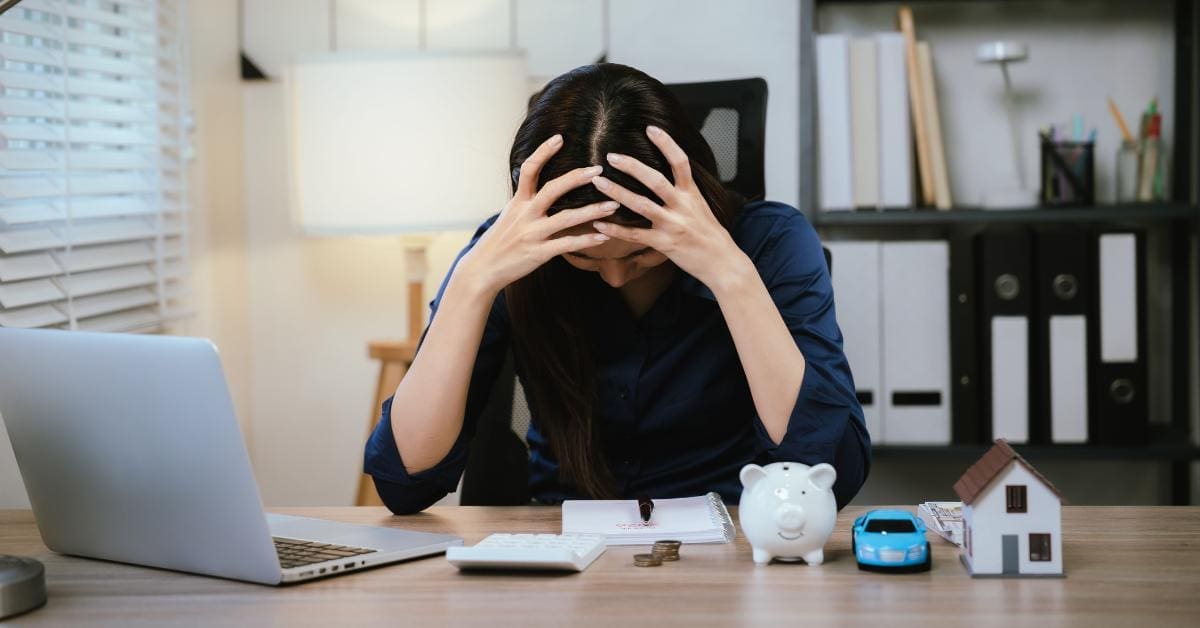 A woman sits, head in hands, over a notepad, laptop, and calculator. Beside her is a piggy bank and a toy car.