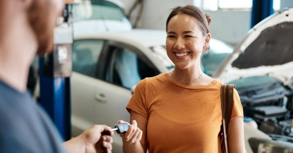A smiling woman in an orange top receives car keys from a mechanic in a garage, with vehicles and workshop equipment visible in the background.