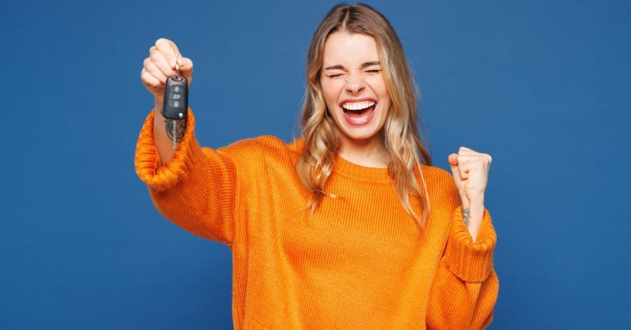 What the new BNPL rules mean for drivers (UK): A smiling woman in an orange jumper celebrating with a clenched fist while holding up a car key, against a blue background.