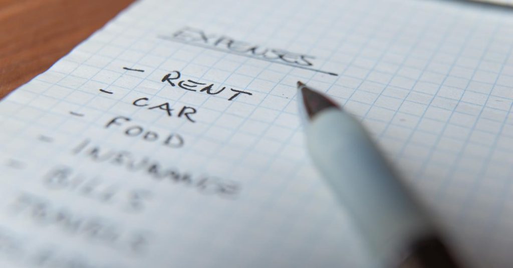 budgeting for car maintenance cost of living A close-up of a handwritten expenses list on squared paper, with the words "Rent", "Car" and "Food" clearly visible, and a pen resting across the page.