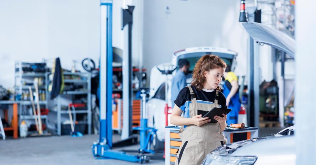 do customers choose garages based on payment options UK A female mechanic in overalls consults a tablet whilst leaning over a car bonnet in a busy garage workshop, with a colleague working on another vehicle in the background.