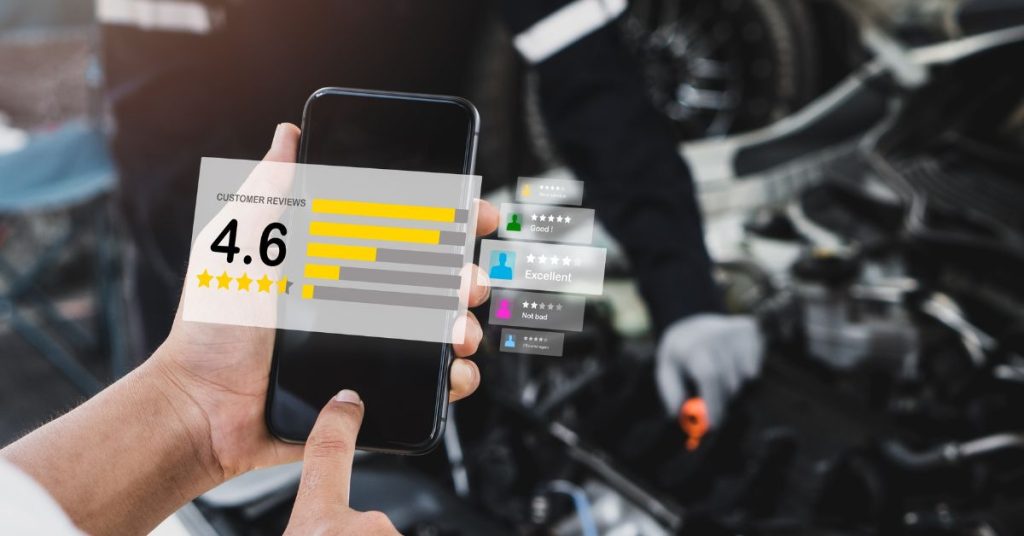 How garages build reputation: A hand holding a smartphone displaying a customer reviews screen with a 4.6 star rating and various review ratings, set against a blurred background of a mechanic working on a car engine.