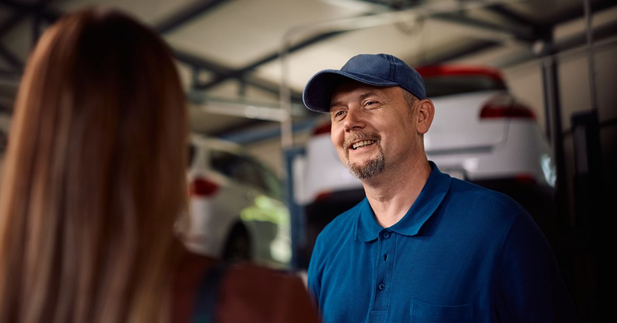 What Makes a Customer Recommend Your Garage: A smiling mechanic in a blue polo shirt and cap talking to a customer whose back is to the camera, in a garage workshop with cars in the background.
